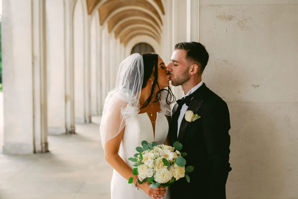 The bride and groom share a tender kiss beneath a magnificent arched colonnade while holding a lush white and green bouquet.