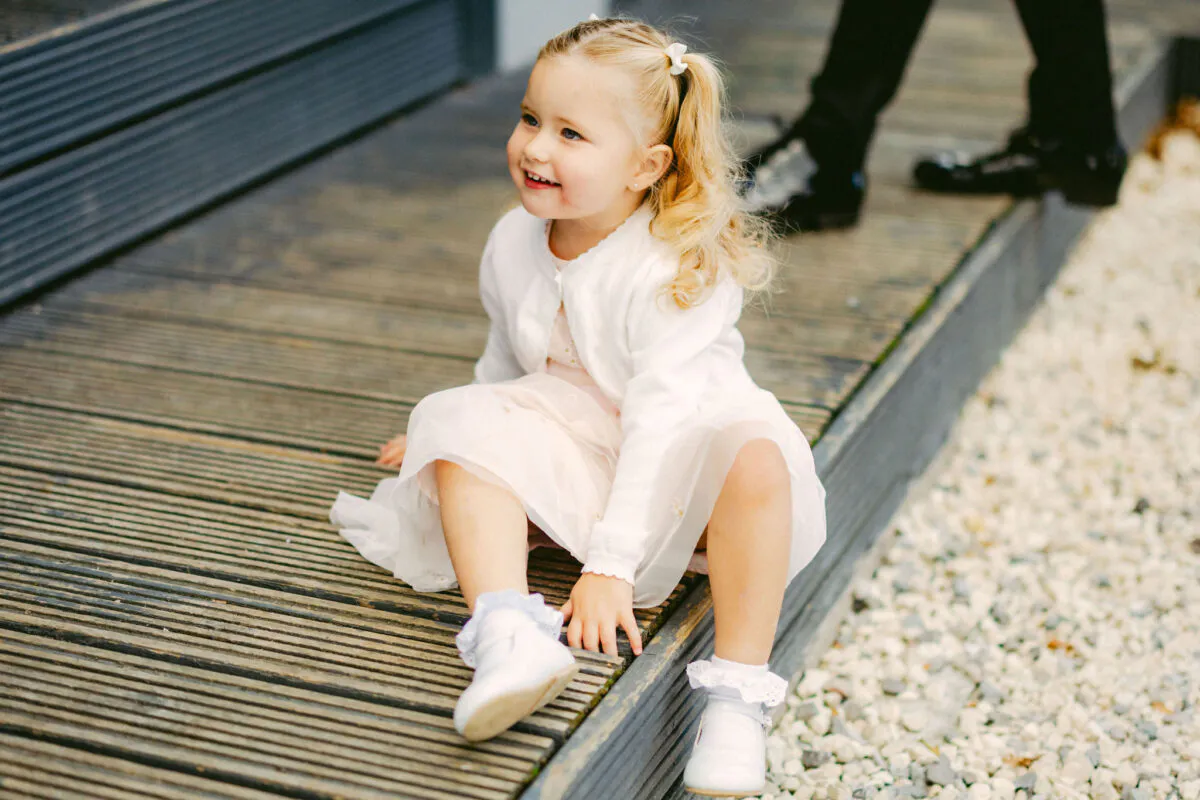 A young flower girl in a white dress and cardigan sits on a wooden deck, smiling candidly during a wedding.