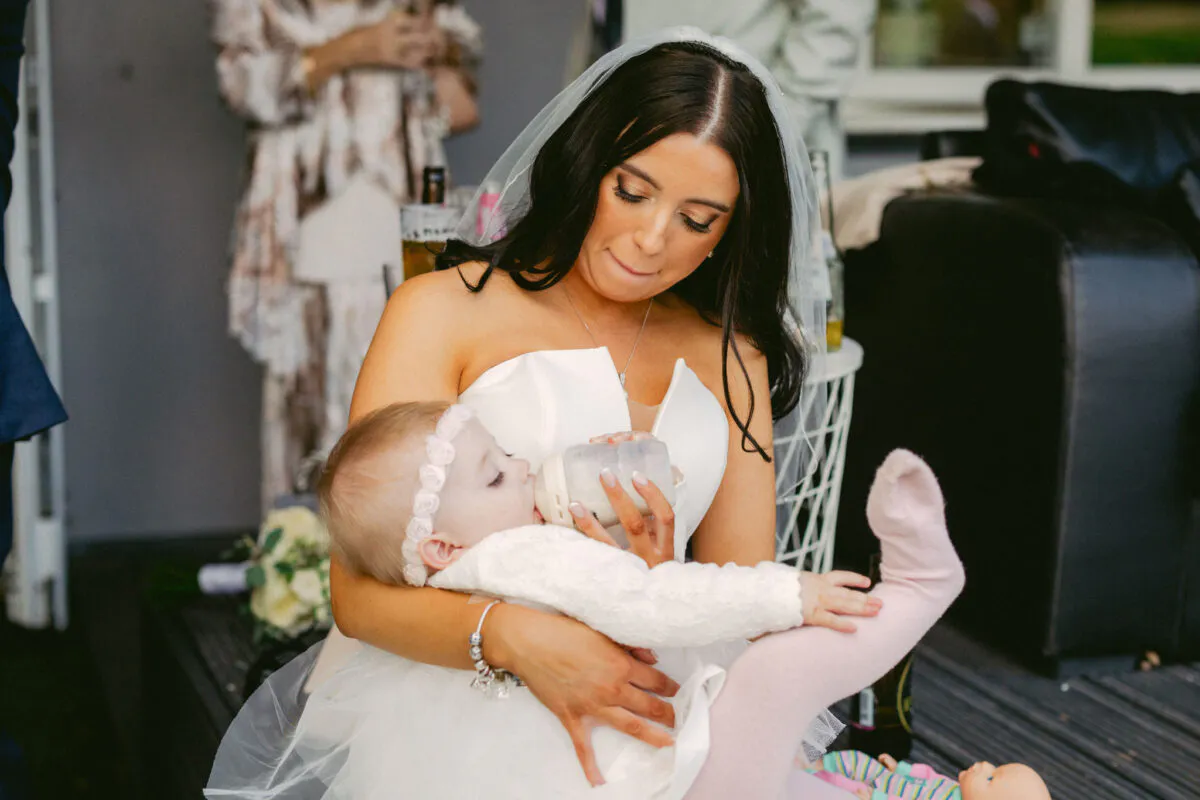 A smiling bride wearing a white strapless gown and veil tenderly holds a baby drinking from a bottle during the wedding celebration.
