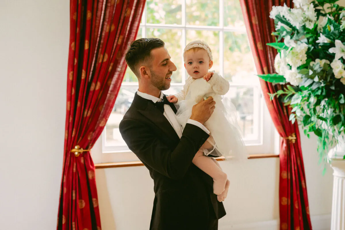 A groom in a classic black tuxedo smiling while holding a baby dressed in white near a window with red curtains and large floral arrangements.