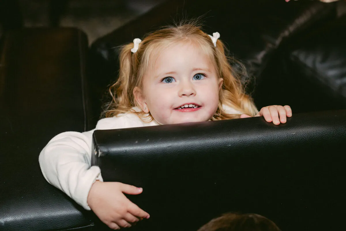 A young flower girl with blonde hair in pigtails smiles happily while peering over the back of a black leather sofa.