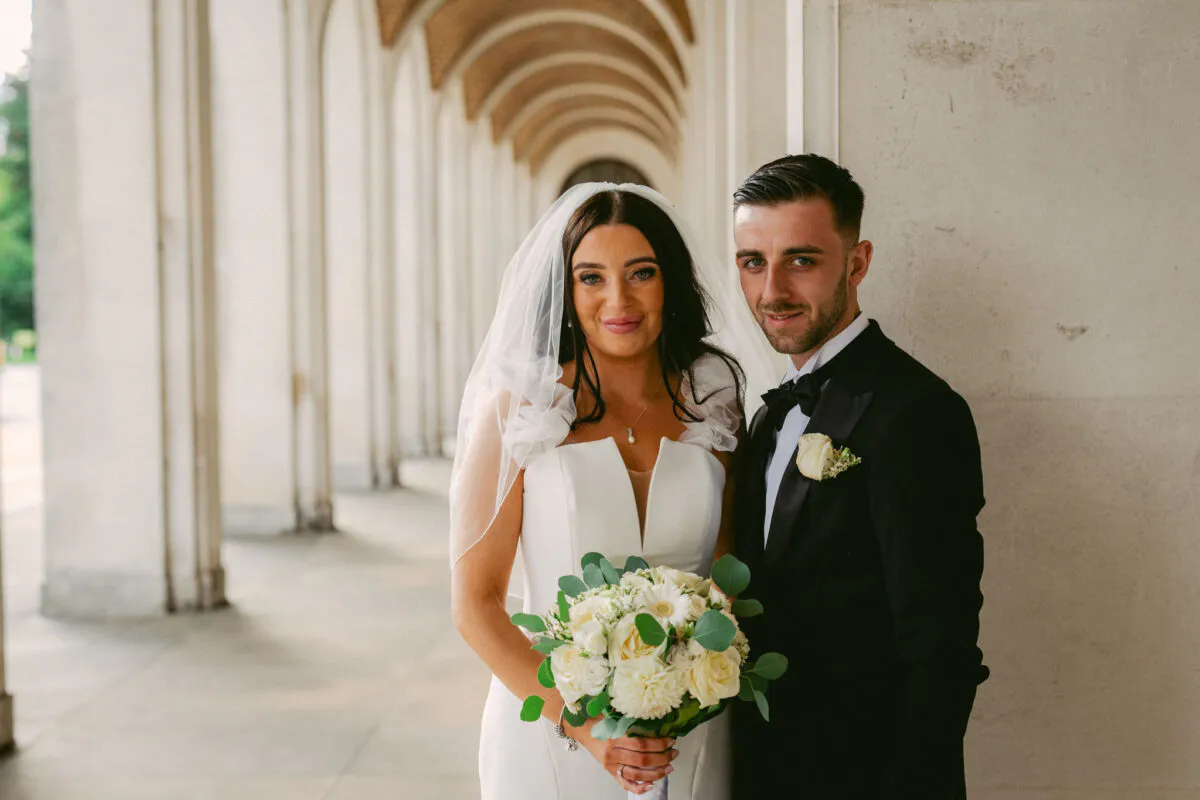 A newly married couple poses gracefully beneath a grand stone archway, with the bride holding a classic white and green floral bouquet.