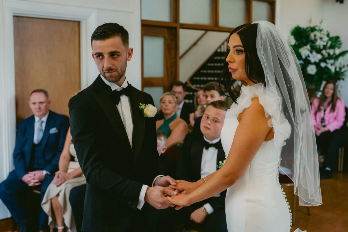 The bride and groom hold hands during their emotional wedding ceremony, with the groom wearing a black tuxedo and the bride in a high-necked white gown and veil.