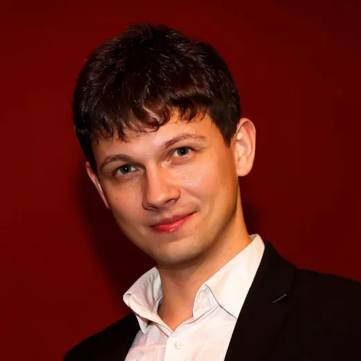 A close up portrait of a groom wearing a dark jacket and a white open collar shirt against a dark red backdrop.