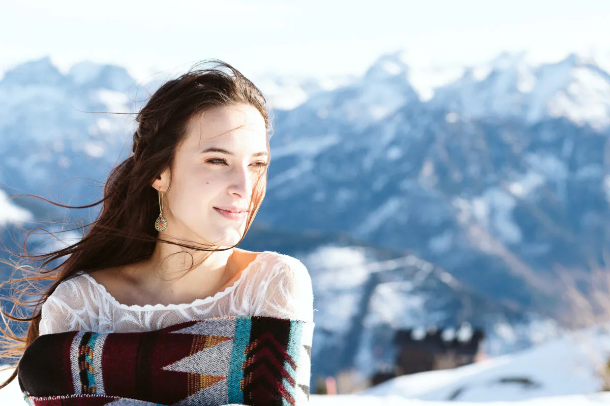 A smiling bride wrapped in a patterned blanket with her hair blowing in the wind against a snowy mountain backdrop.