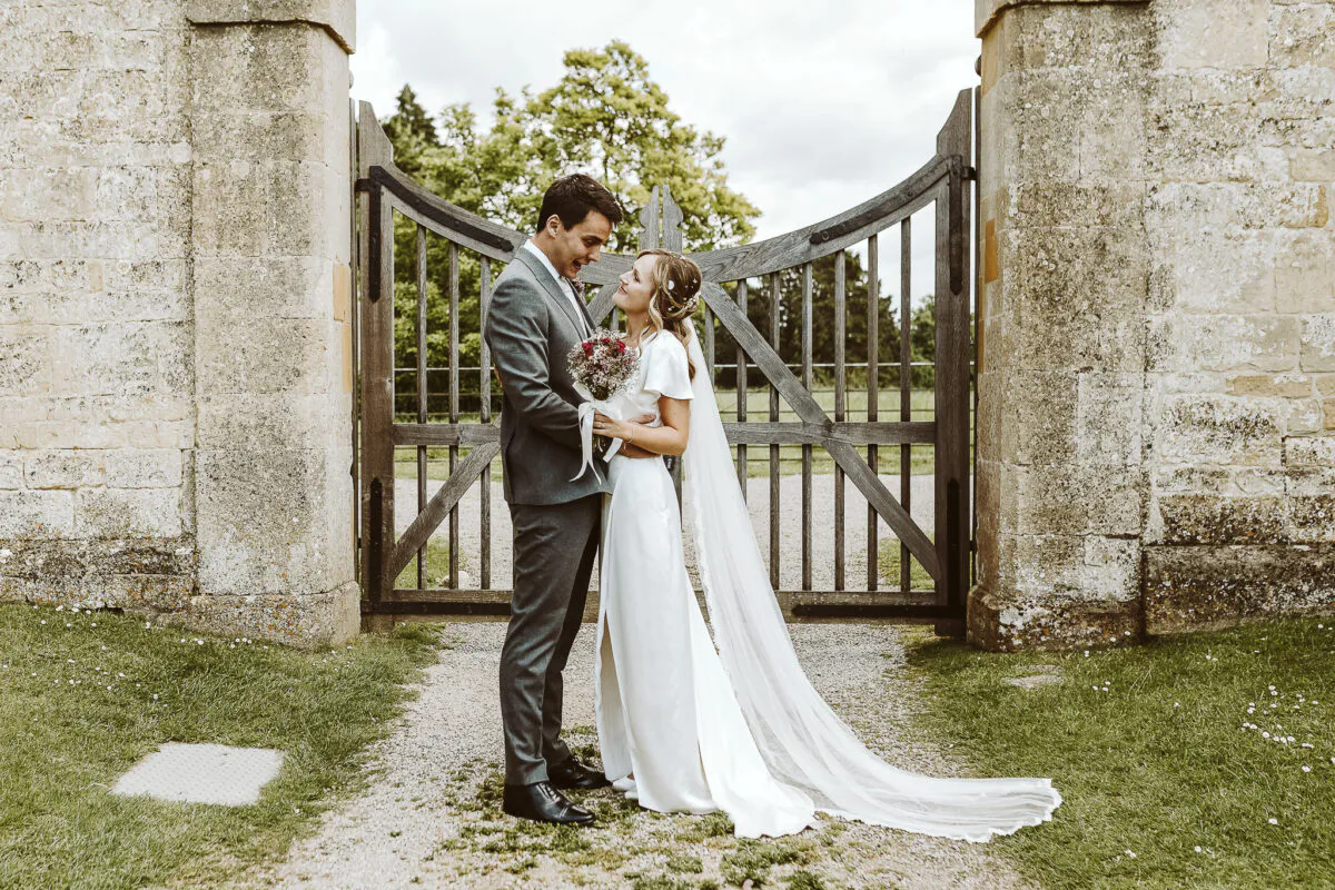 A bride and groom stand lovingly outside grand wooden gates flanked by stone pillars, with the bride holding a delicate bouquet and wearing a flowing veil.