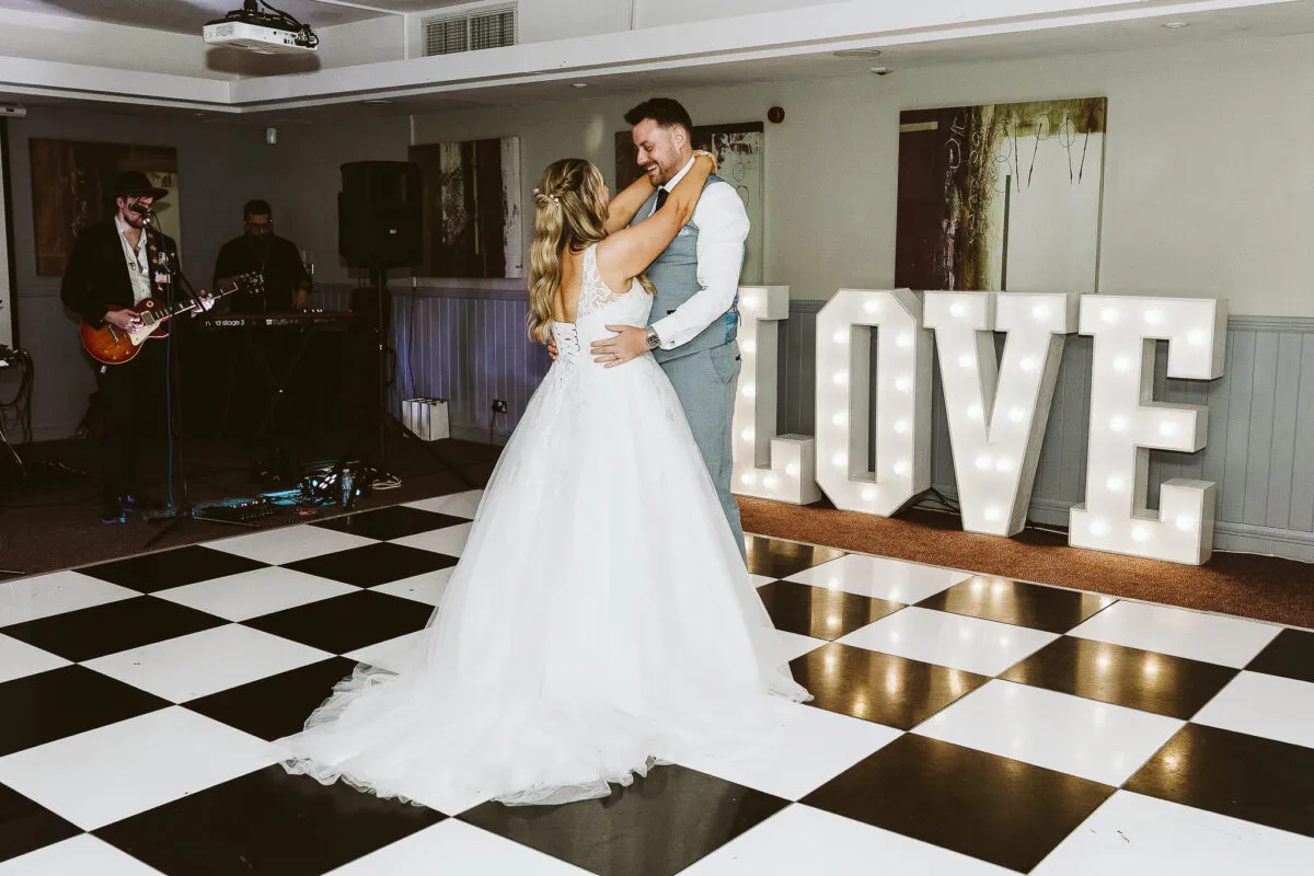 Newlyweds share their first dance on a black and white checkerboard dance floor while a live band plays and large illuminated letters spell out love in the background.