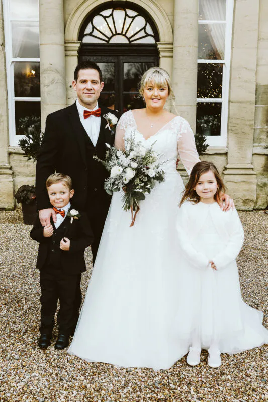 A bride and groom pose outside a grand stone manor house with a young page boy and flower girl, holding a lush white and greenery bouquet.