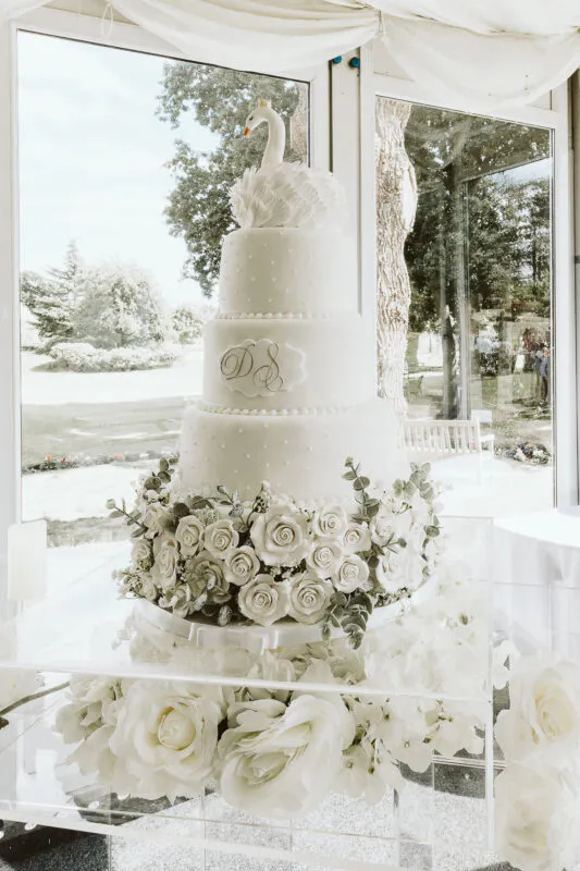 An elegant tall tiered white wedding cake featuring a monogram plaque and a delicate swan topper rests on a clear acrylic stand surrounded by cascading white roses.