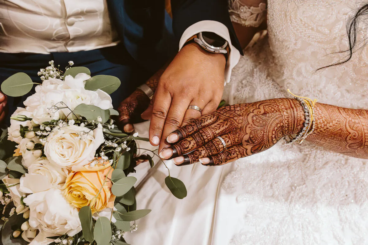 Close-up of a newlywed couple holding hands displaying wedding bands, detailed henna patterns on the bride's hand, and a bouquet of white roses.