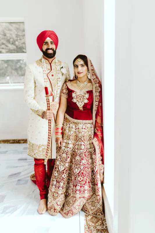 A newlywed Sikh couple poses together indoors, wearing traditional embroidered attire including a red bridal lehenga and a cream sherwani with a red turban.