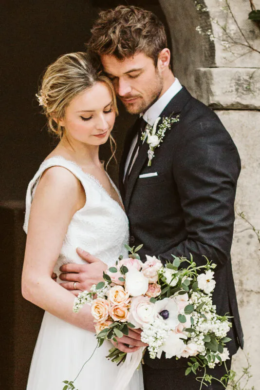 A newlywed couple embraces tenderly in front of a stone archway, with the groom resting his head against the bride while holding a large pastel floral bouquet.