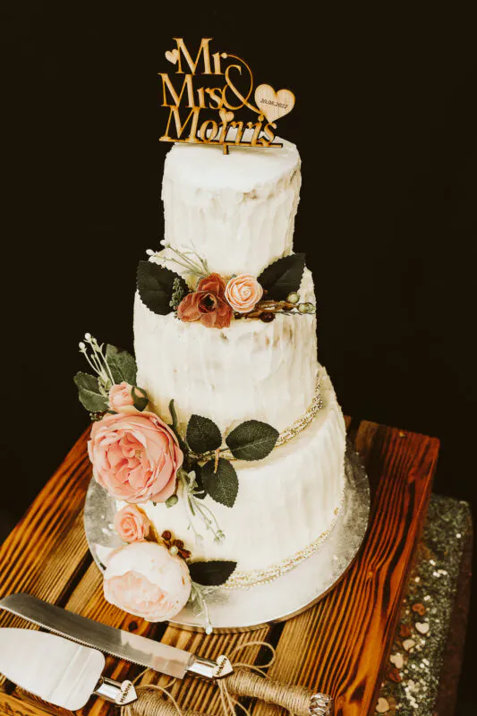 A three-tier semi-naked wedding cake adorned with pink and peach roses and a wooden Mr and Mrs cake topper rests on a rustic wooden stand.