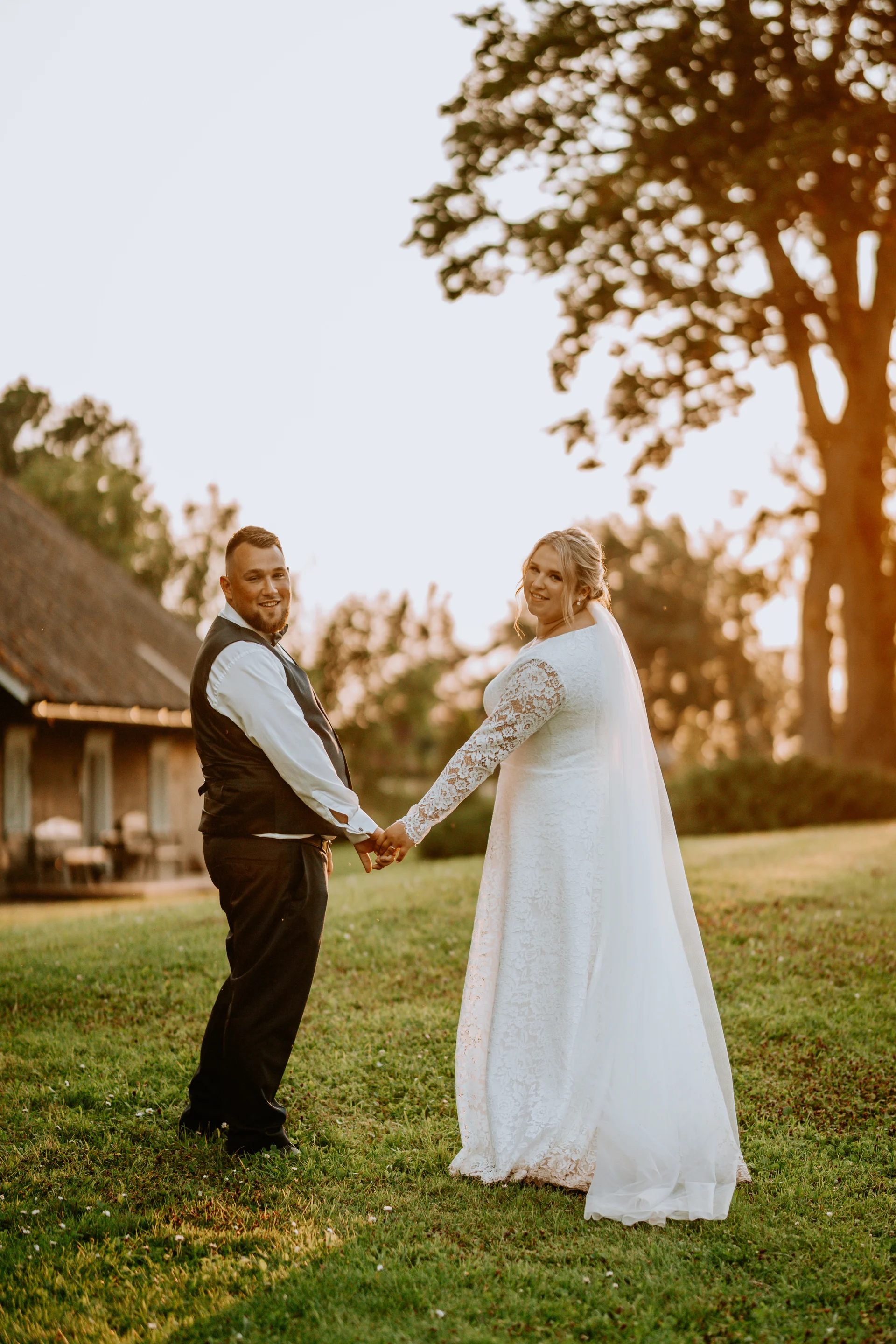 A bride and groom hold hands and look back smiling during sunset on their wedding day, standing on a grassy hill near a rustic countryside building.