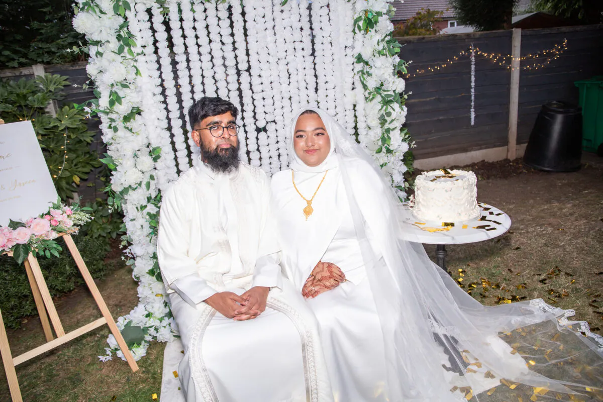 A newly married couple sits together in front of a white floral backdrop during their outdoor nikah ceremony, with a decorated cake on a small table beside them.