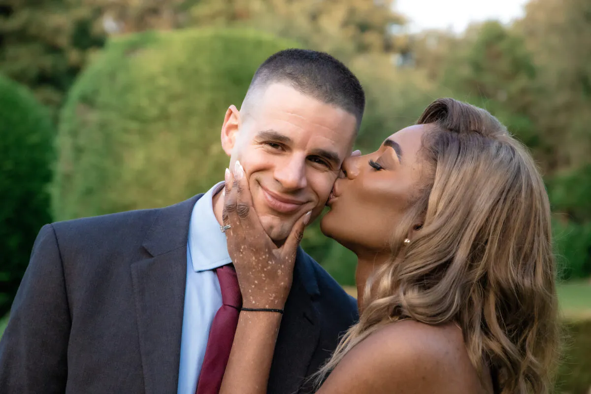 An interracial couple shares a tender moment outdoors as the bride kisses the groom's cheek while holding his face with both hands.