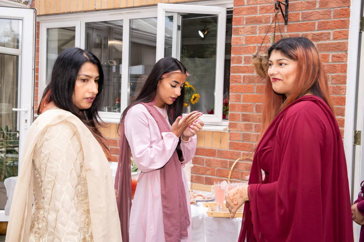 Three guests in traditional attire gather outside during a wedding celebration, with henna visible on their hands and soft natural light illuminating the scene.