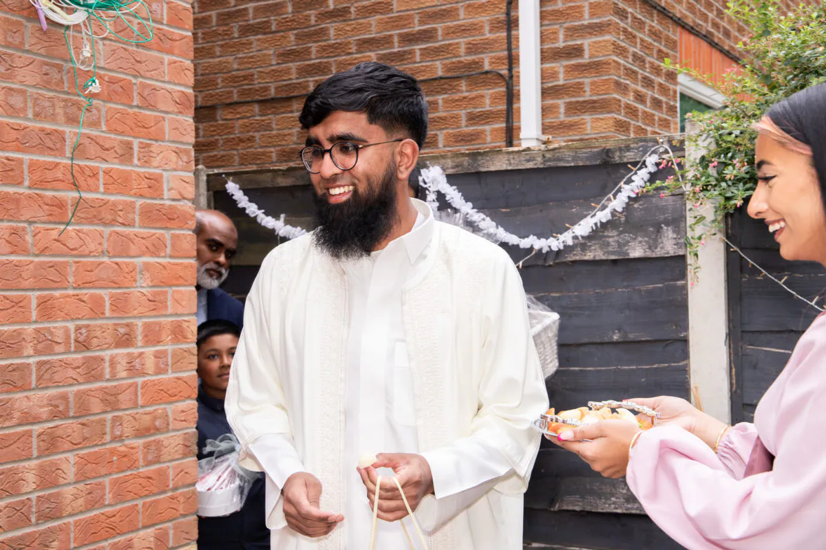 A smiling groom wearing a traditional white kaftan stands outdoors near a brick wall during a wedding celebration.