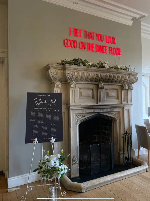 An elegant wedding seating plan rests on a white easel next to an ornate stone fireplace adorned with floral arrangements and a bright red neon sign.