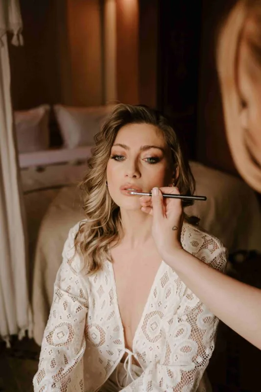 A close-up photograph captures a bride having her lipstick applied by a makeup artist while wearing a white lace robe during morning preparations.