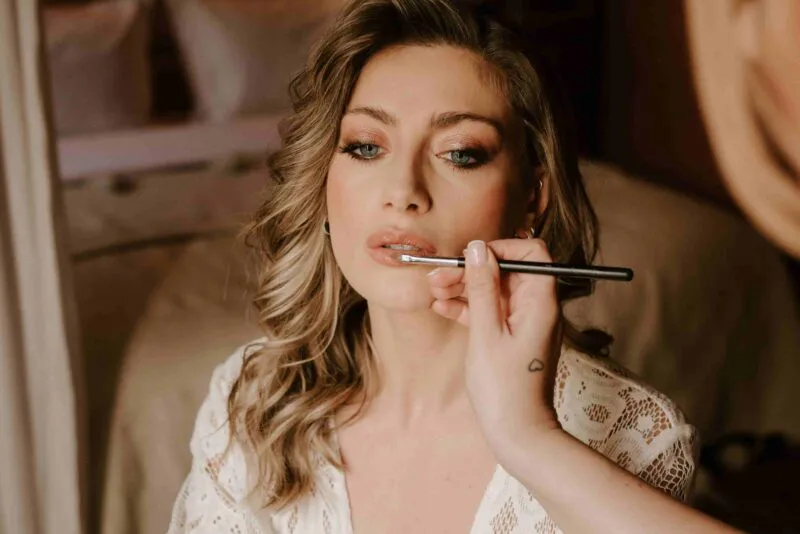 A close-up photograph captures a bride having her lipstick applied by a makeup artist while wearing a white lace robe during morning preparations.