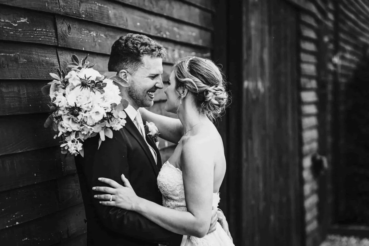 A newlywed couple shares a joyful moment laughing nose to nose in front of a rustic wooden barn while the bride holds a textured bouquet.