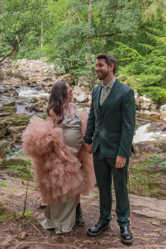 A pregnant bride wearing an olive silk slip dress with a dramatic ruffled pink tulle wrap smiles adoringly at her groom, who wears a smart forest green suit, beside a rushing river in a lush woodland setting.