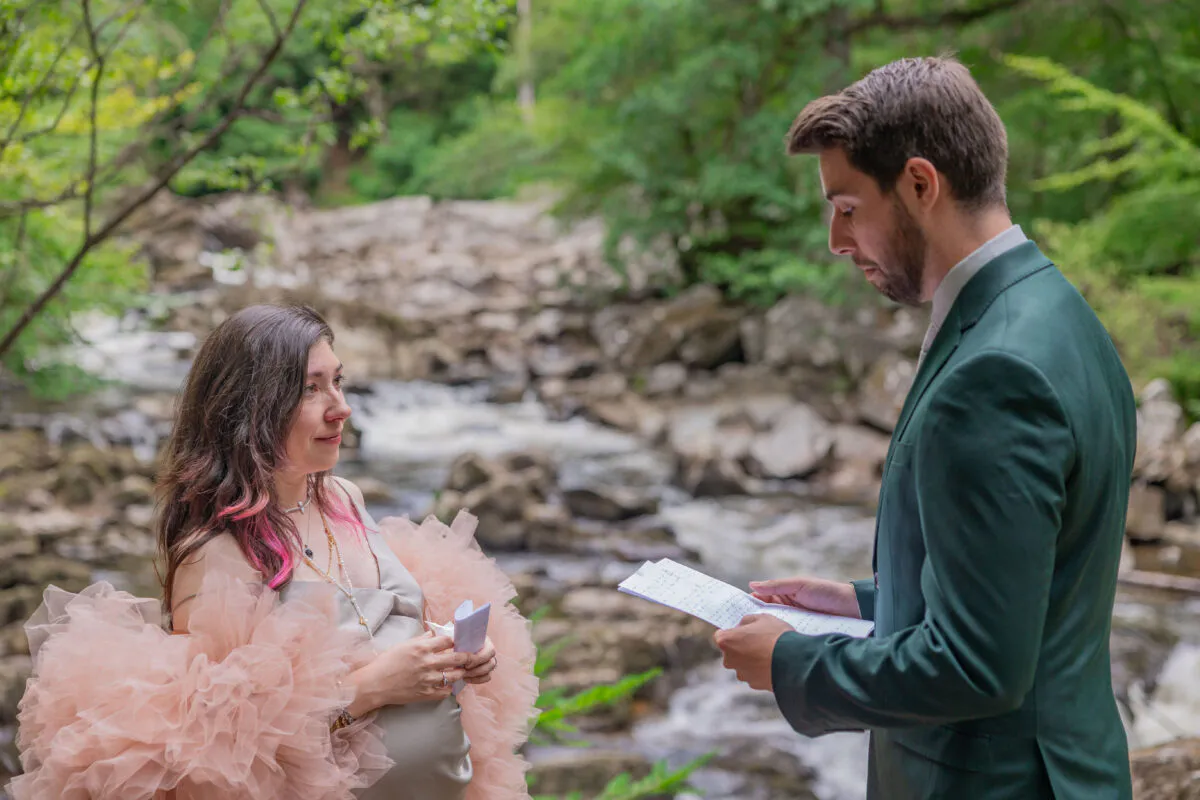 A couple exchanges personal vows during an outdoor elopement ceremony beside a rocky river in a lush woodland setting.