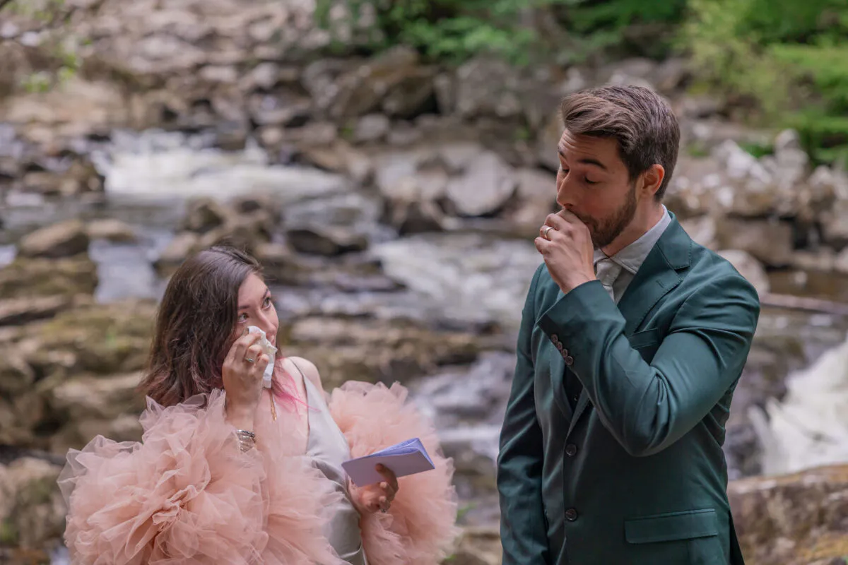 An emotional couple shares a tender moment reading vows outdoors by a rocky river, with the bride wiping away tears and the groom looking touched.