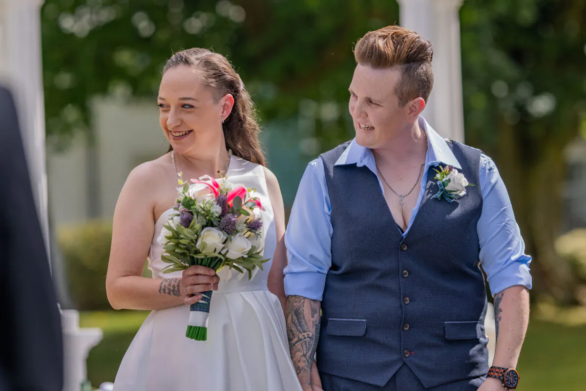 Two smiling brides stand outdoors during their wedding ceremony, one holding a hand-tied bouquet of roses and thistles while wearing a white dress, and the other wearing a blue shirt and waistcoat.