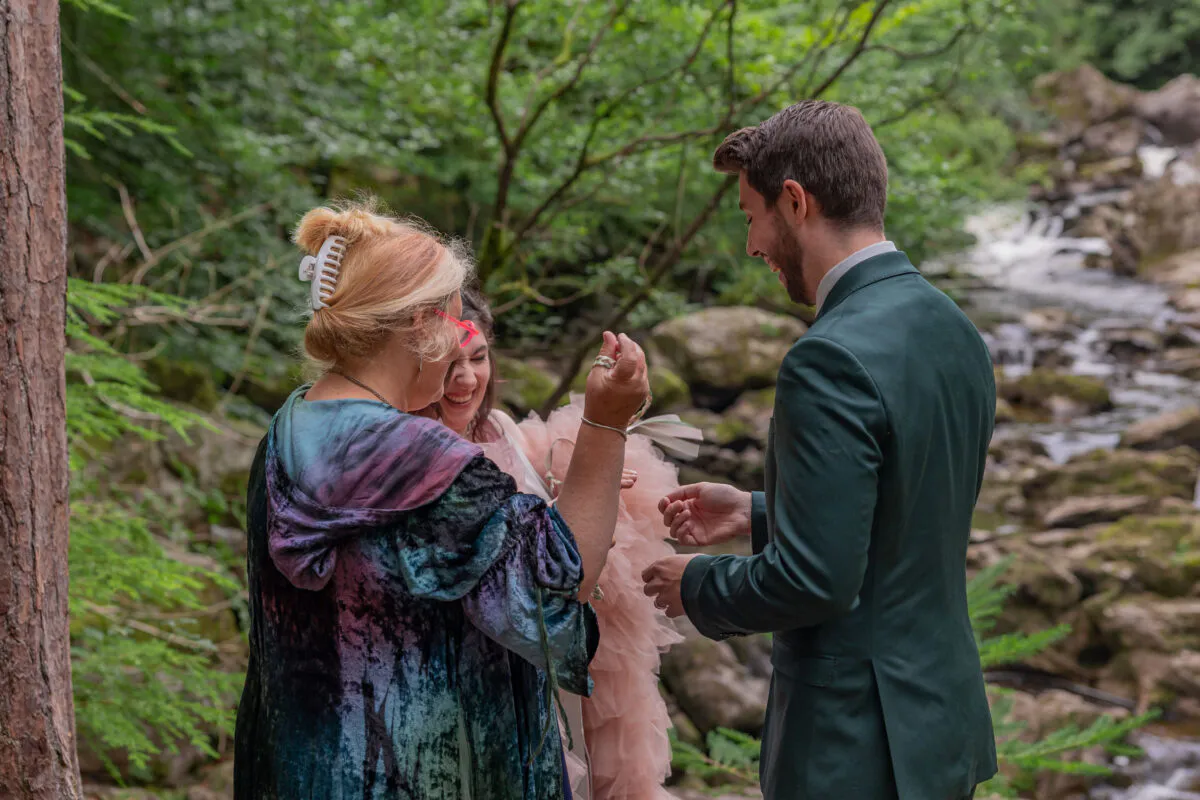 A celebrant conducts a handfasting ceremony for a couple in a lush woodland setting beside a flowing river.