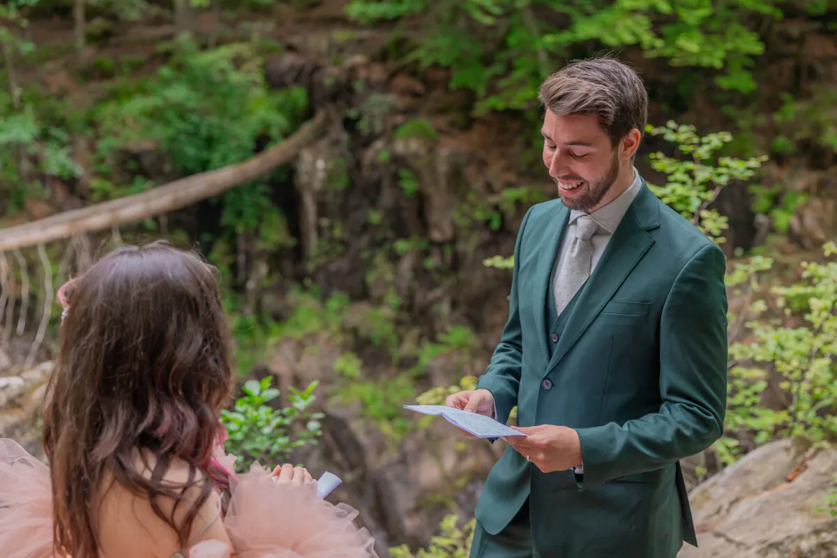 A groom in a green suit smiling as he reads vows from a card during an outdoor woodland ceremony with his partner.