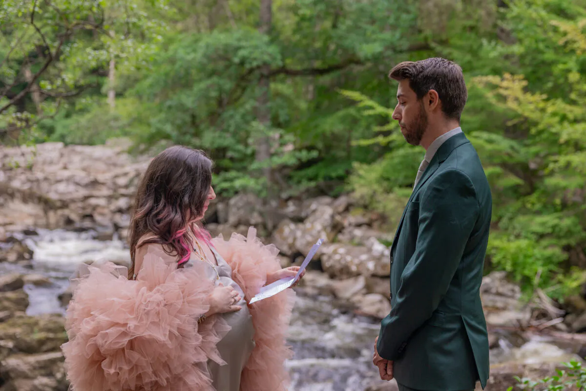 A pregnant bride wearing a dramatic pink tulle dress reads vows from a letter to a groom in a dark green suit during an intimate woodland ceremony by a rushing river.