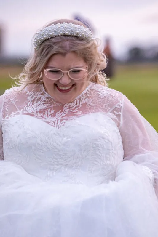 A smiling bride wearing a lace wedding dress with an illusion neckline and a pearl headband laughs happily outdoors in soft light.