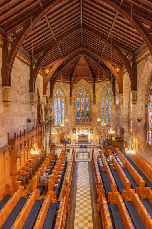 An elevated wide-angle view of a grand historic church interior, featuring high vaulted wooden timber ceilings, tall stained glass windows, and rows of polished wooden pews lining a chequered aisle.