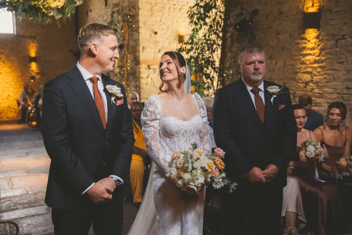 A smiling bride holding a floral bouquet shares a joyful glance with her groom inside a rustic stone barn during the wedding ceremony.
