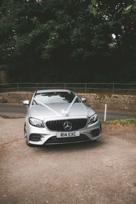 A silver modern wedding car is decorated with white ribbons across the bonnet, parked on a gravel driveway against a backdrop of trees and a stone wall.