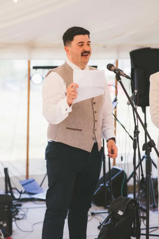 A groom delivers a speech with a smile while holding notes inside a marquee during the wedding reception.