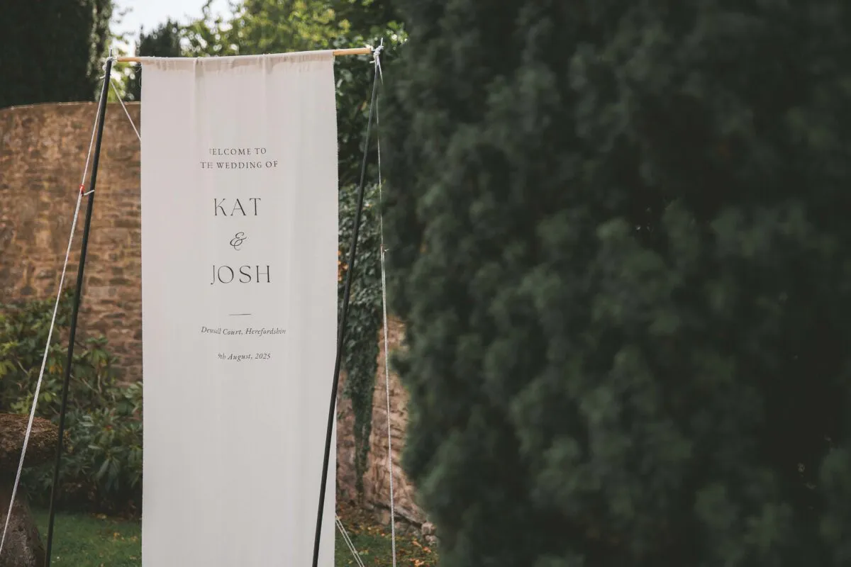 A modern fabric welcome sign featuring minimalist typography stands outdoors against a stone wall and lush greenery.