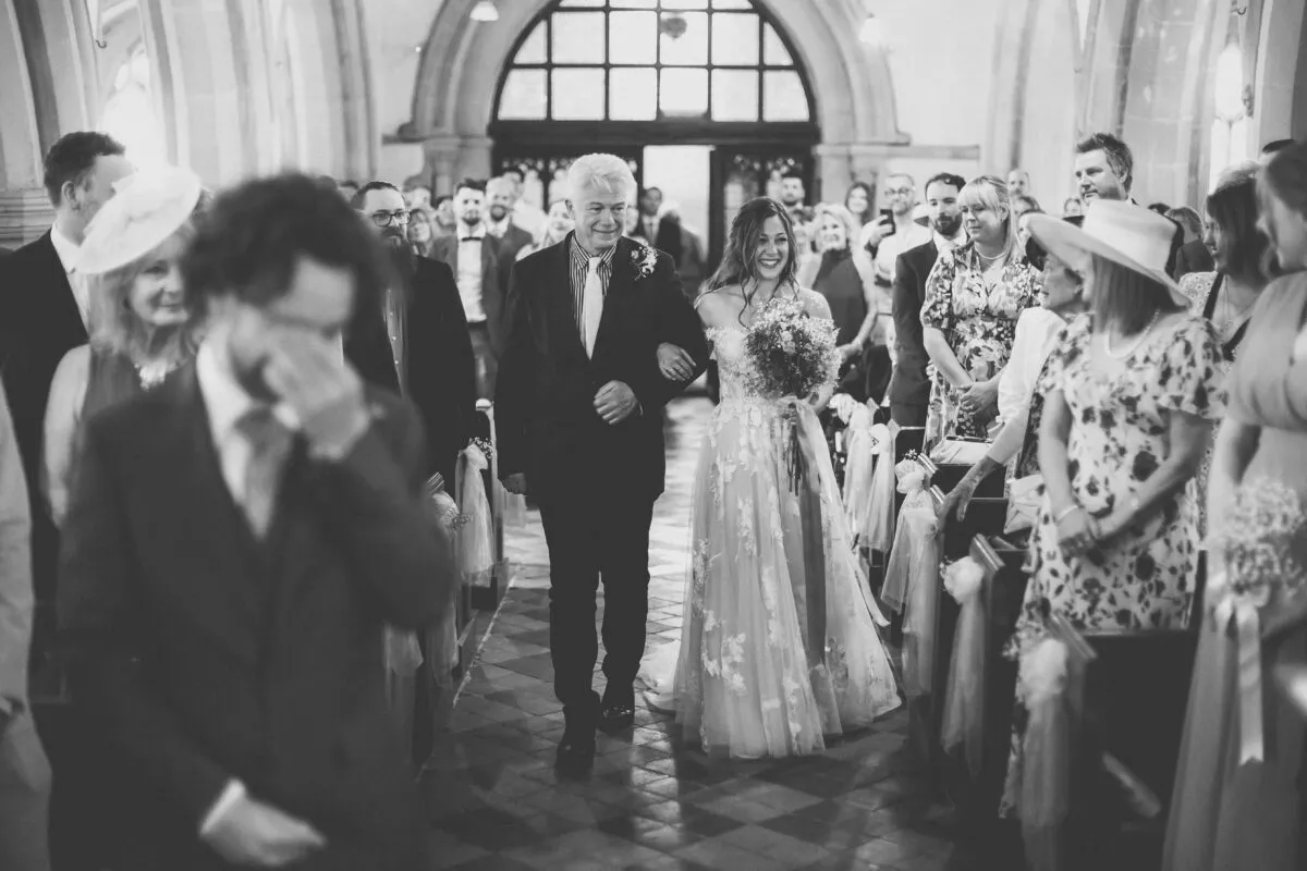 A bride smiling warmly while walking down the church aisle arm in arm with her father, watched by an emotional congregation.