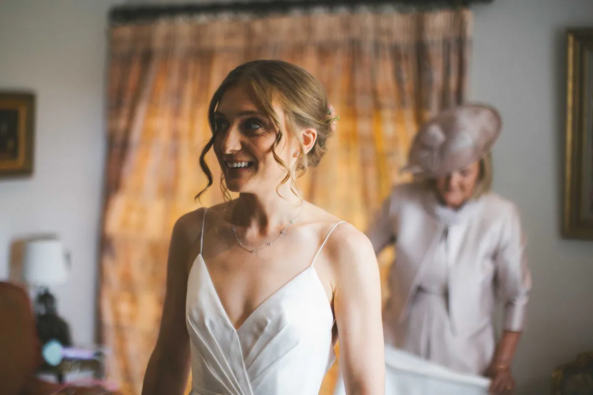 A joyful bride in a simple slip dress smiles while getting ready indoors, with her mother looking on in the background.