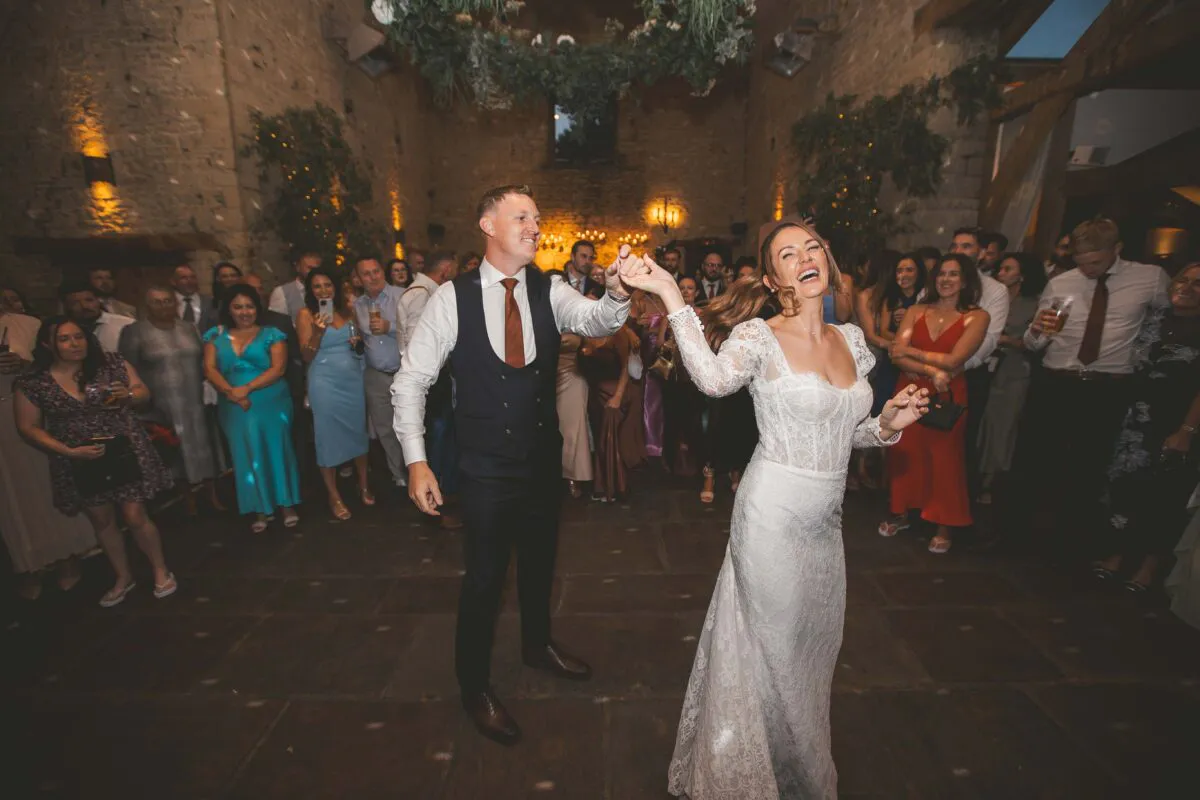 A joyous bride and groom share their first dance surrounded by cheering wedding guests in a rustic stone barn with a hanging floral installation.