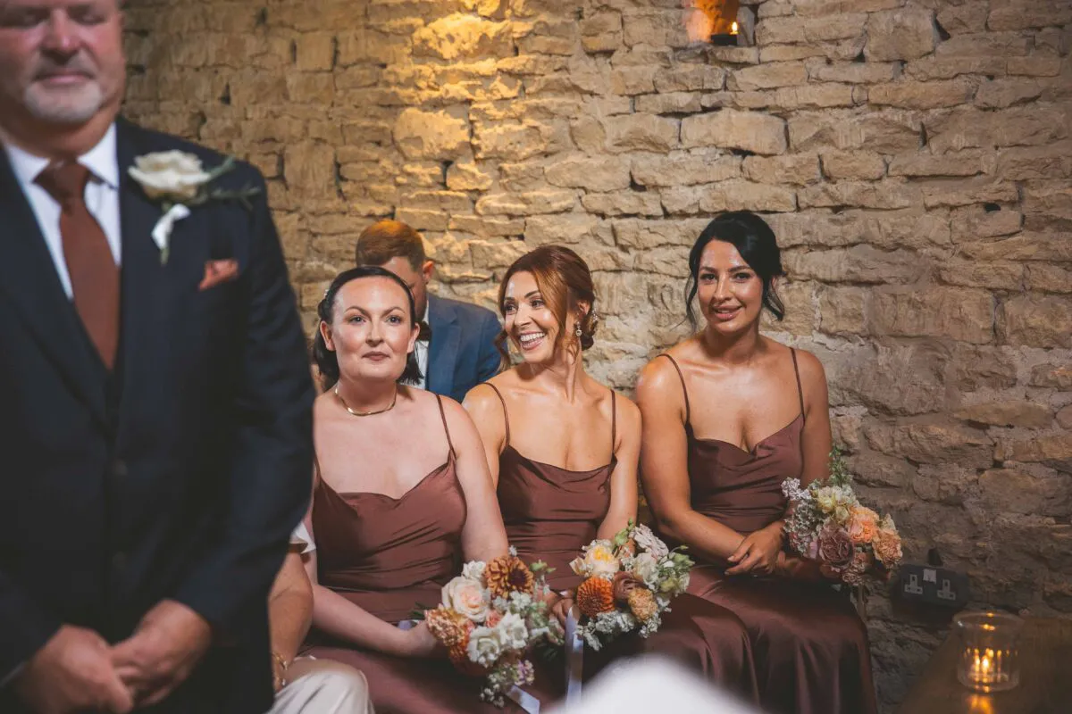 Three smiling bridesmaids in matching satin brown dresses holding floral bouquets are seated together inside a rustic stone barn venue during a wedding ceremony.