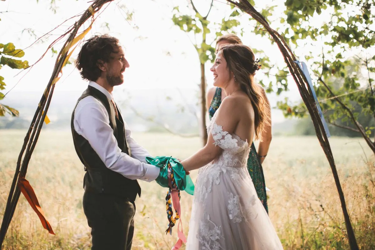 A bride and groom smile at each other during an outdoor handfasting ceremony beneath a rustic wooden arch decorated with colourful ribbons in a meadow.