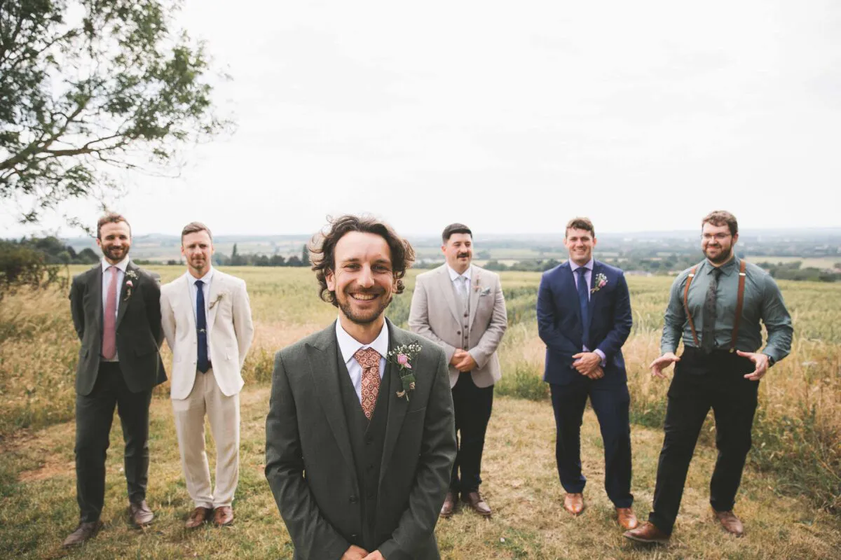 The groom stands proudly in the foreground wearing a green three-piece suit and patterned tie, while his groomsmen in mismatched suits smile in a rural countryside setting.