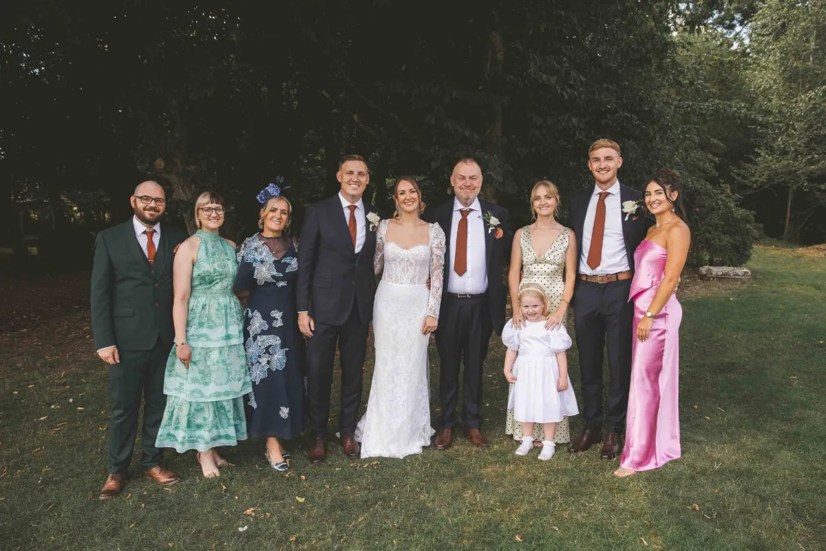 A smiling bridal party and family members stand together on a lush green lawn outdoors, dressed in suits, floral dresses, and formal evening wear.