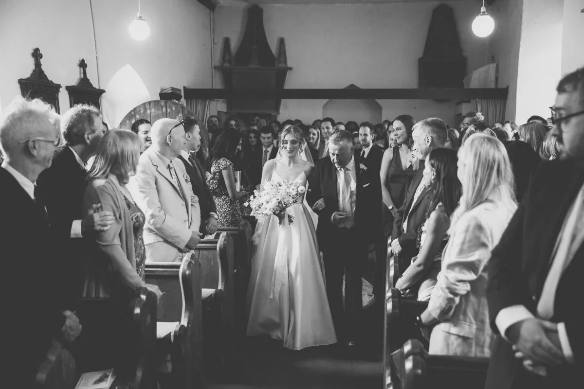 A bride walks down the church aisle holding her father's arm and carrying a bouquet, surrounded by seated wedding guests.