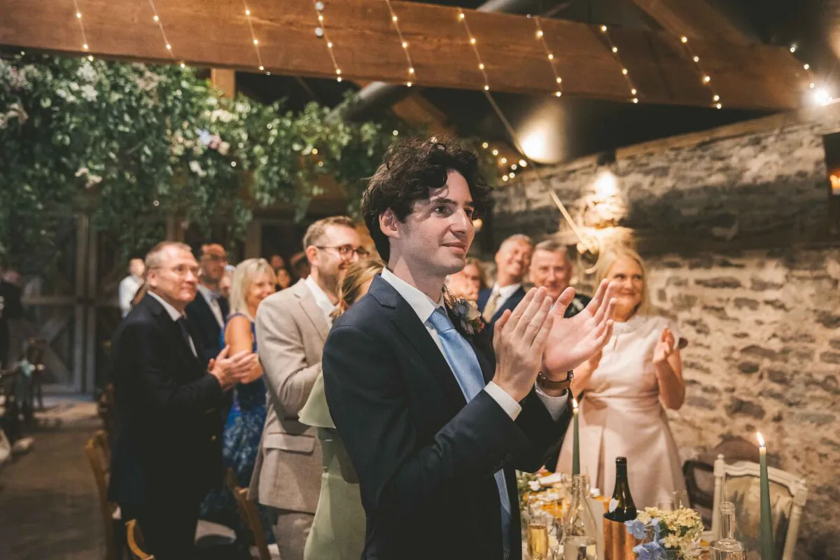 A groom in a navy suit and light blue tie enthusiastically applauds during the wedding reception inside a rustic stone barn adorned with fairy lights.