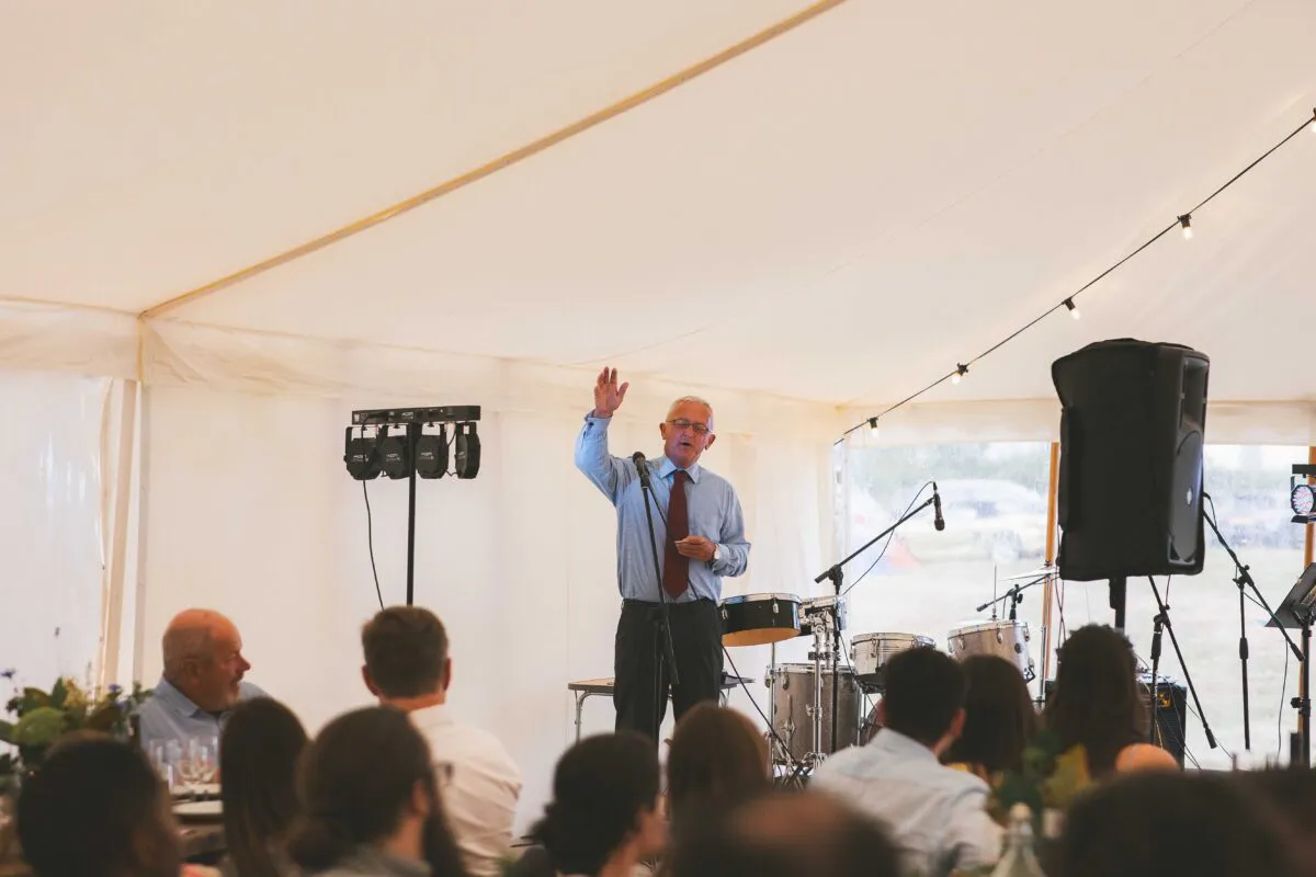 An older man wearing glasses and a red tie delivers a speech with his hand raised inside a bright marquee wedding reception.