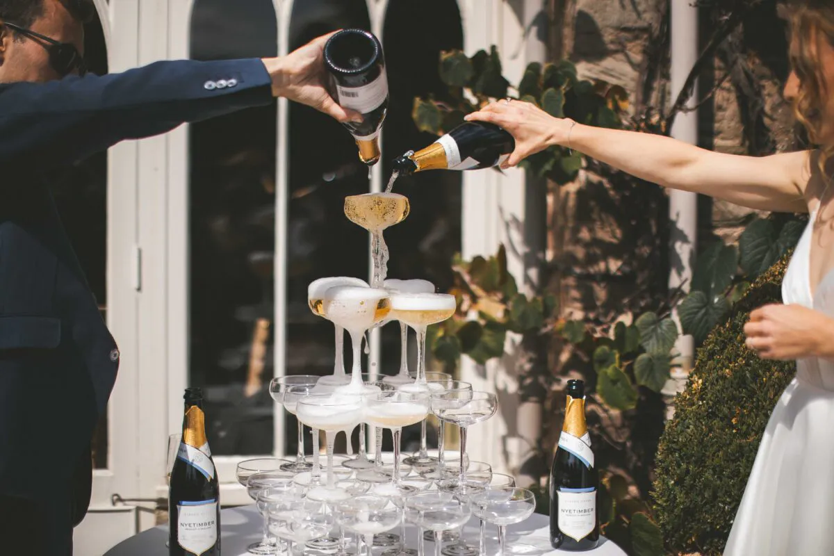 A bride and groom joyfully pour champagne down a multi-tiered coupe tower outside in the bright sunshine.
